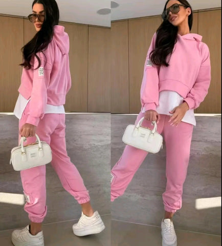 Woman wearing pink hoodie and joggers with white sneakers and sunglasses, holding a white handbag, standing in front of a