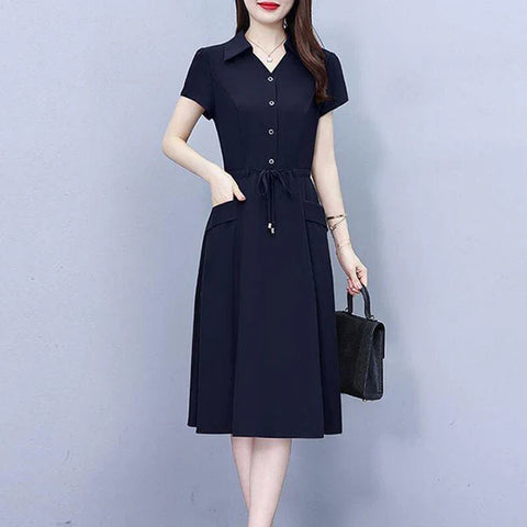 Woman wearing navy short-sleeve button-up dress with drawstring waist and midi length skirt, holding black handbag, standing