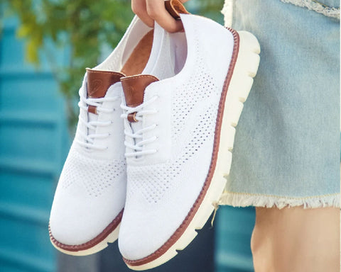 White perforated lace-up shoe with cognac brown trim and cream sole, held in hand against blurred blue water background.