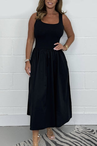 Woman wearing sleeveless black midi dress with gathered skirt, standing against white brick wall with hand on hip.