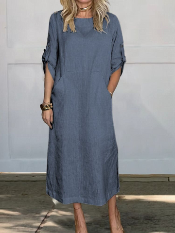 Woman wearing a blue-gray linen dress with rolled three-quarter sleeves, standing against a white wall with hands in pockets.