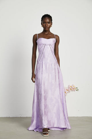 Woman wearing lavender spaghetti-strap maxi dress with draped bodice and floral pattern, holding pastel flower bouquet