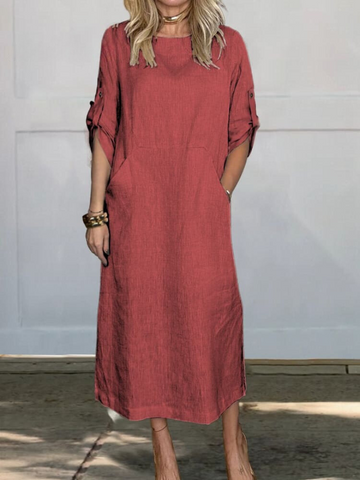 Woman wearing a rust-red linen shift dress with rolled three-quarter sleeves, standing against a white paneled wall. She