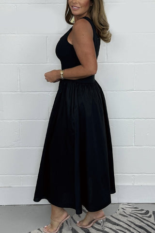 Woman in black sleeveless dress with full skirt standing against white brick wall, wearing gold bracelet and strappy