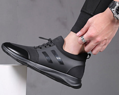 Person wearing black lace-up sneaker with textured upper and thick black sole, paired with black sweater and watch, foot