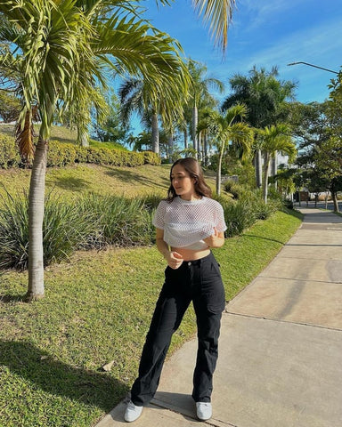 Woman standing on sidewalk wearing white crop top and black pants, surrounded by palm trees and green lawn under blue sky.