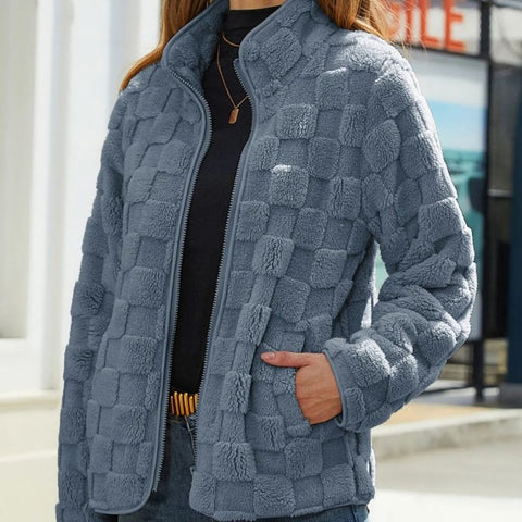 Woman wearing a blue-gray quilted zip-up jacket with a checkerboard pattern over a black top, standing outdoors with hand in