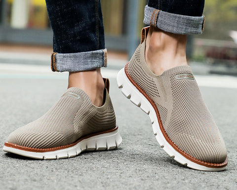 Person wearing rolled dark jeans standing on pavement in beige knit slip-on sneakers with white soles and brown trim.