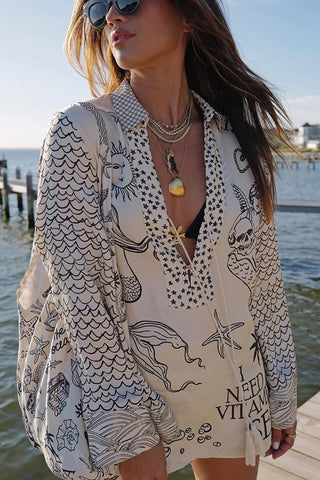 Woman wearing oversized white button-up shirt with black line drawings of fish and ocean motifs, sunglasses, and layered
