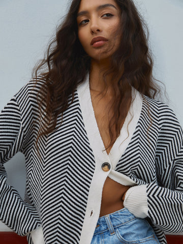 Woman wearing black and white striped cardigan over white crop top with blue jeans, looking downward against light gray