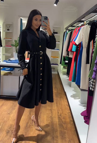 Woman in black button-front dress with belt taking mirror selfie in bright retail clothing store with wooden floor and