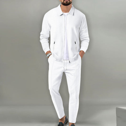 Man wearing white zip-up jacket and matching white pants, hands in pockets, standing against gray background.