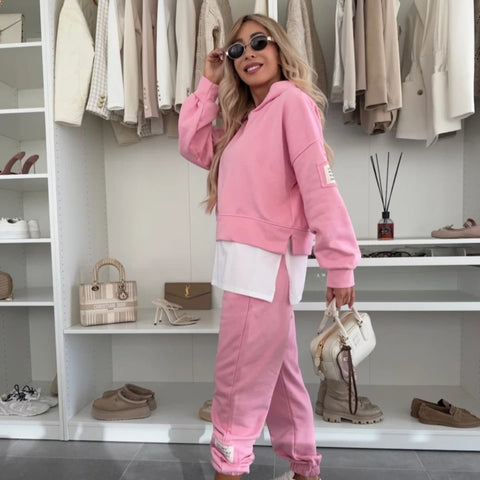 Woman wearing pink sweatsuit with white stripe detail, dark sunglasses, standing in closet with neutral-toned clothing and