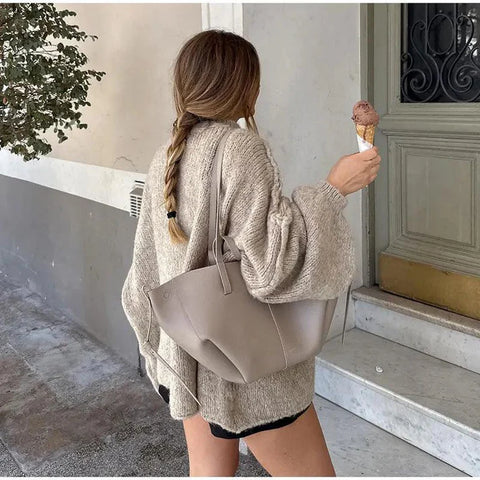 Woman from behind wearing beige knit cardigan over black shorts, carrying light tan tote bag, holding ice cream cone