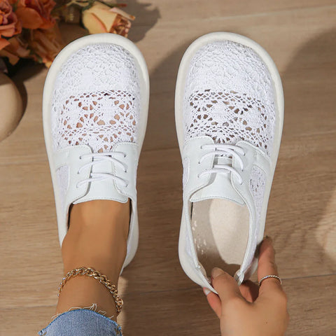 White lace-panel sneakers with perforated details, laced closure, worn on feet against wooden floor with flowers visible in