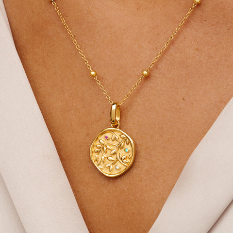 Gold chain necklace with circular pendant featuring raised leaf design, worn on warm-toned skin against white clothing.