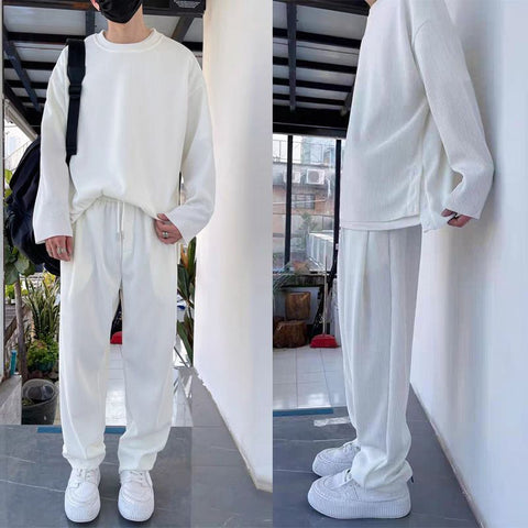 Person wearing white crew neck sweatshirt and white pants with white sneakers, standing indoors near windows and plants