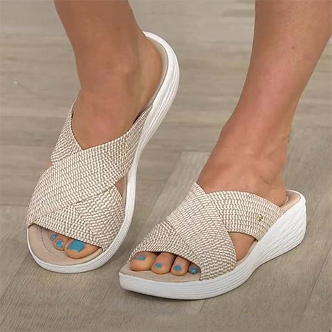Pair of beige textured slide sandals with white soles, open-toe design, worn on feet with blue nail polish against wooden
