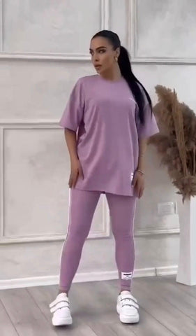 Woman wearing lavender oversized t-shirt and matching leggings with white sneakers, standing in minimalist interior with