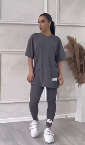 Woman wearing dark gray oversized t-shirt and black leggings with white sneakers, standing against white paneled wall with