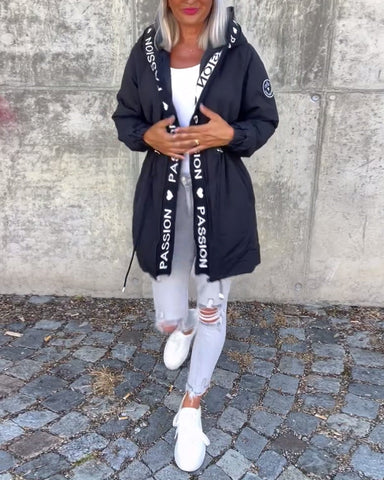 Woman wearing black hooded puffer jacket over white top, white distressed jeans, and white sneakers, standing against