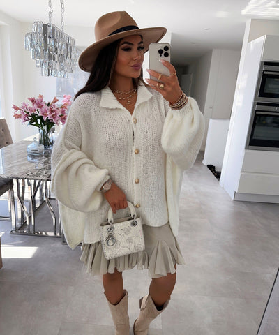 Woman in cream cardigan and pleated skirt holding phone to face, wearing brown hat and white boots, standing in modern
