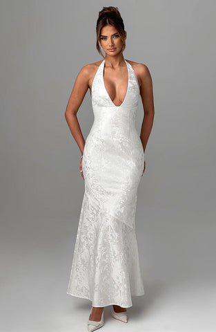 Woman wearing white halter neckline gown with lace texture and fitted bodice, standing against neutral gray background.