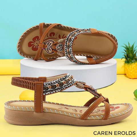 Brown leather sandals with decorative beaded straps and embroidered details, displayed on white platform against turquoise
