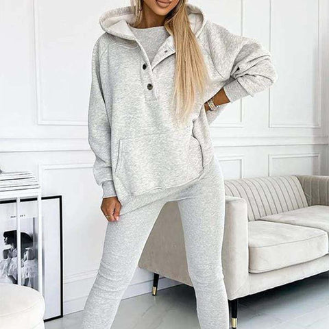 Woman wearing light gray button-front hoodie and matching jogger pants, standing with hand on hip in front of white paneled