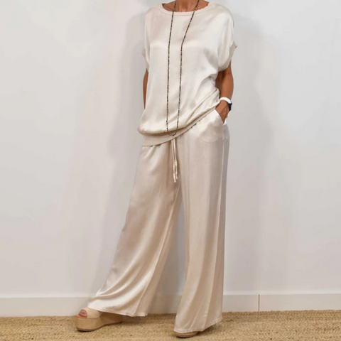 Woman wearing cream short-sleeve top and matching wide-leg pants with drawstring waist, standing against white wall with