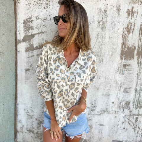 Woman wearing leopard print button-up shirt and distressed denim shorts, sunglasses, standing against weathered white wall.