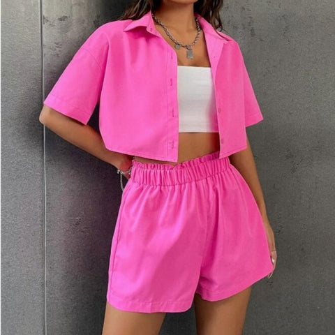 Woman wearing bright pink button-up shirt and matching shorts with white crop top underneath, standing against gray textured