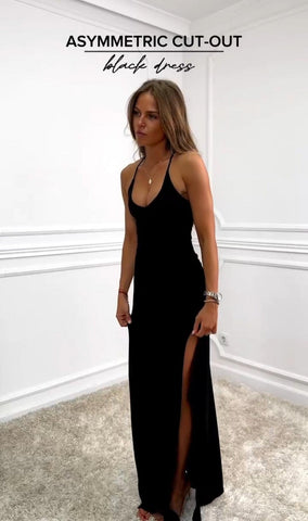 Woman wearing black halter maxi dress with asymmetrical cut-out detail on side, standing against white wall with hand